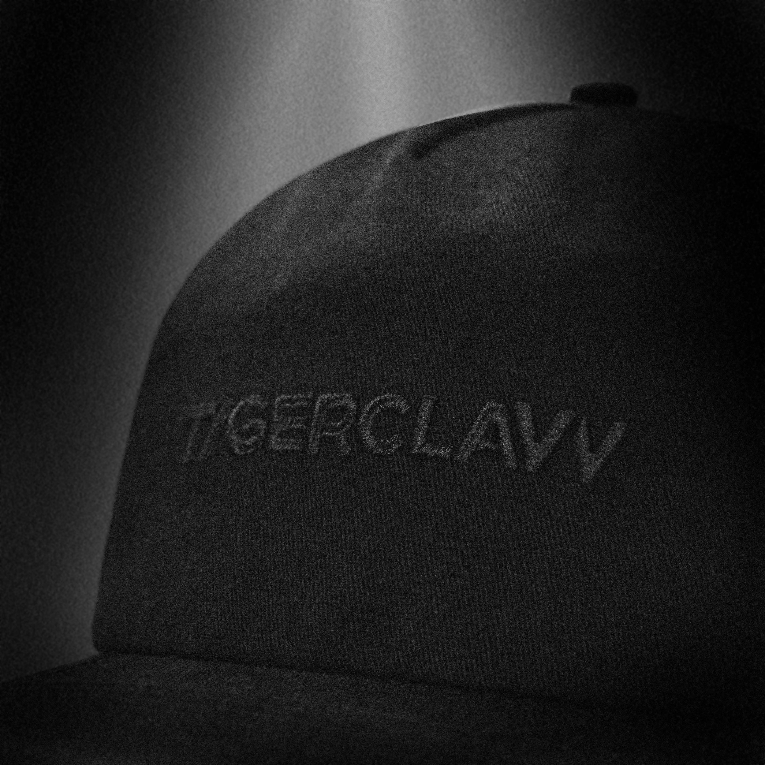 Close-up of the Tigerclaw Black Stash cap in monochrome, highlighting the tonal black-on-black embroidered “TIGERCLAW” logo on the crown with a moody spotlight effect.