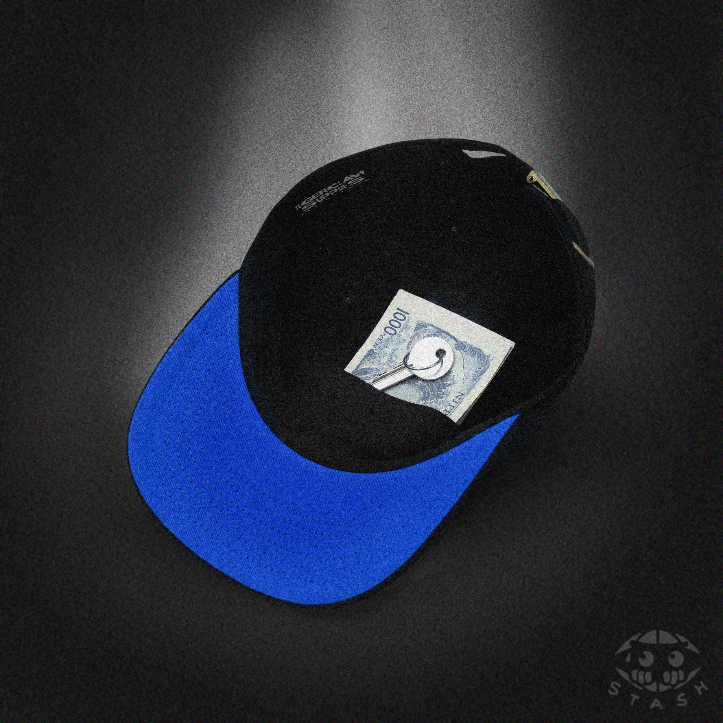 Black Stash cap with bright blue brim shown from an angled underside view, revealing a secret stash pocket holding a folded banknote and key inside the crown, presented on a moody spotlight background with the Stash icon logo.