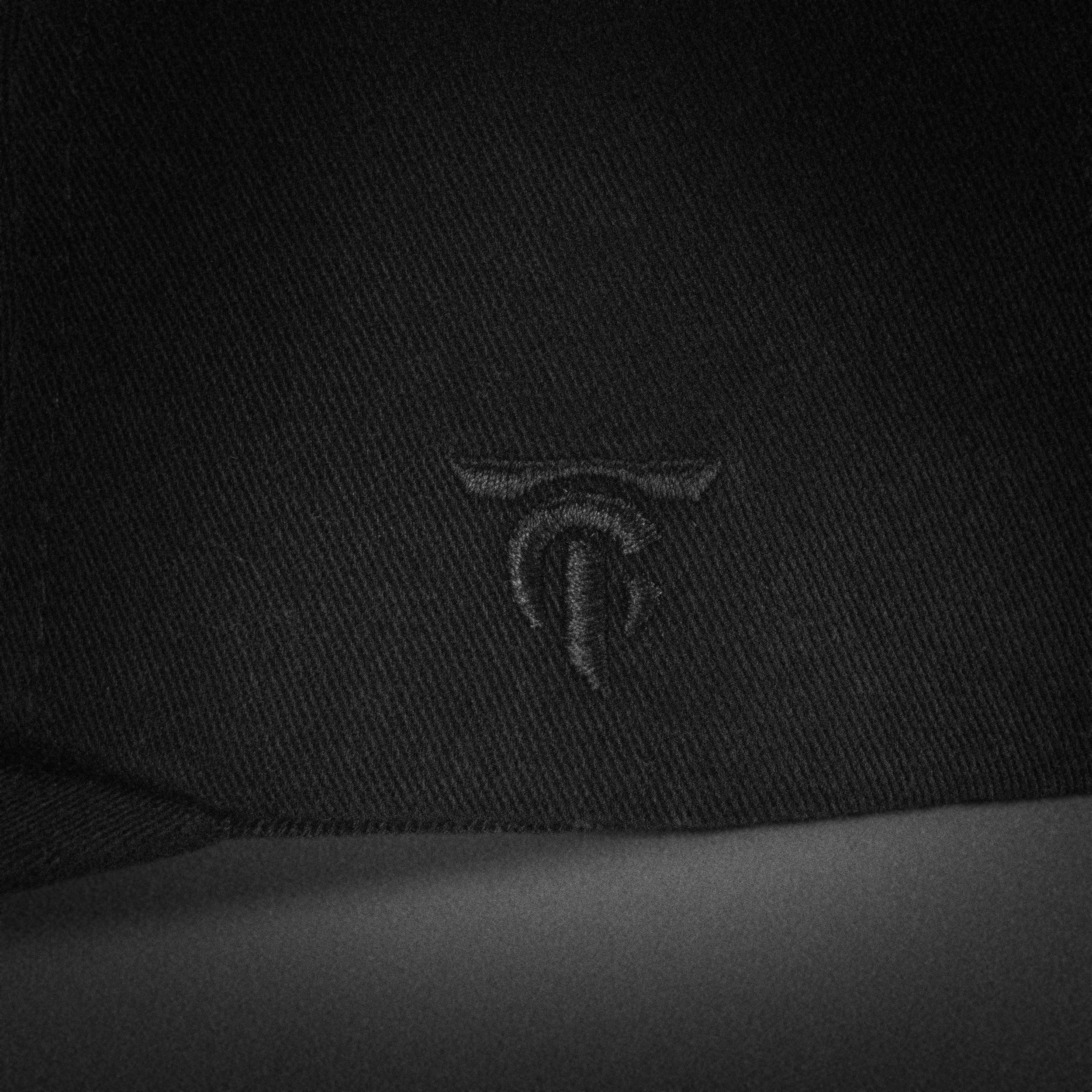 Close-up of the Tigerclaw Black Stash cap showing the tonal black embroidered TC monogram logo on the side panel, shot in a moody monochrome style.