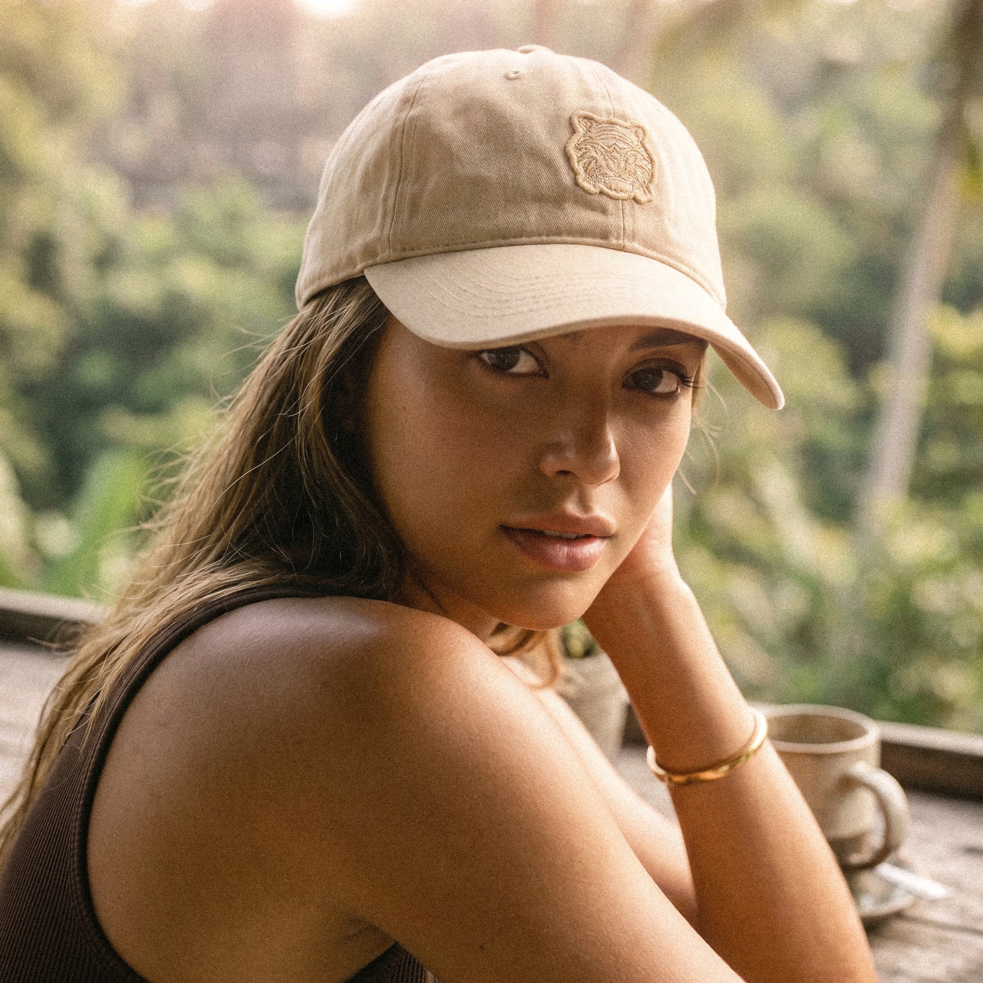 Woman wearing a beige cap with a tigerclaw logo, sitting outdoors with a blurred natural background, Close-up portrait of a person wearing the Tigerclaw Classic Sand Beige Strapback cap, showcasing premium everyday headwear in a neutral sand colorway