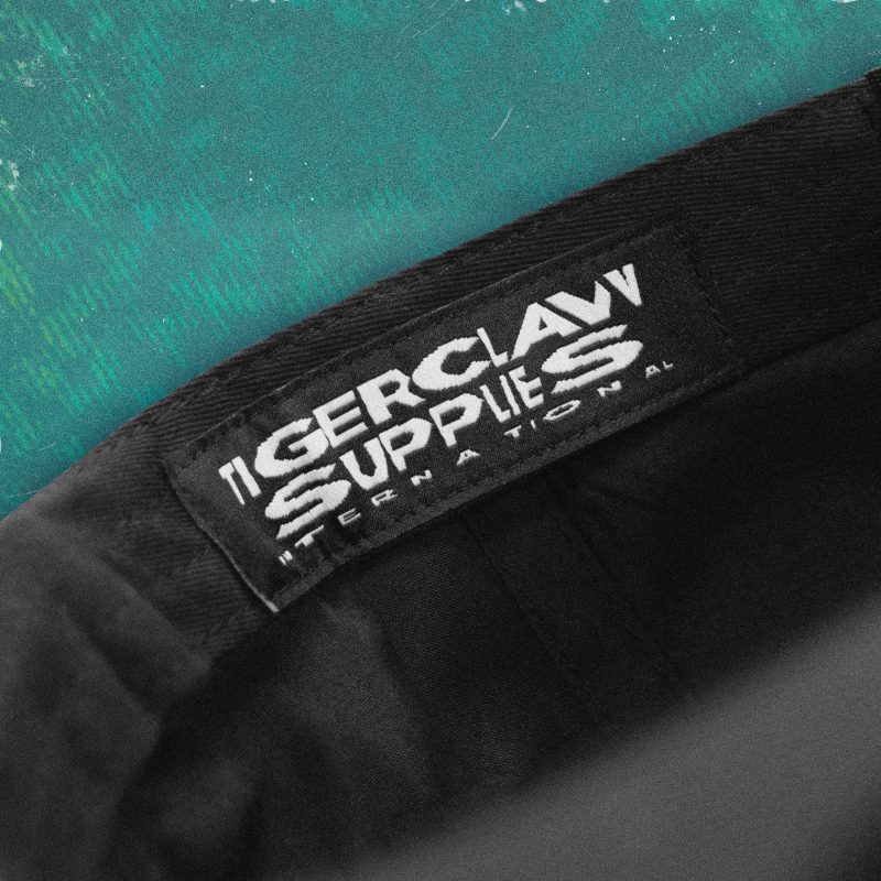 Close-up of inside woven label on black nylon cap reading “TIGERCLAW SUPPLIES INTERNATIONAL” in bold white embroidery, against a teal textured background.